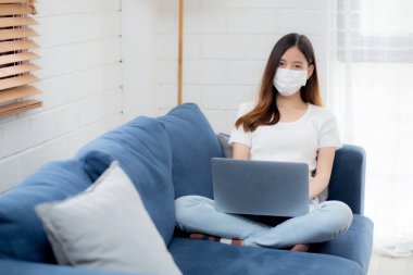 Yüz maskeli Asyalı genç bir iş kadını evde dizüstü bilgisayardan internete, oturma odasında internete, tıbbi maskeli bir kız ve covid-19 salgını için karantinaya alınıyor..