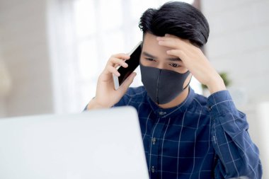 Yüz maskeli Asyalı genç bir iş adamı akıllı telefondan konuşuyor ve bilgisayar başında Coperid-19 'u korumak için çalışıyor. İş adamı karantina işini evden defter ve sosyal mesafeyle yapıyor ve evde kalıyor..
