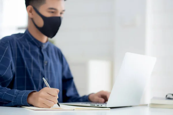 Young asian business man in face mask for protect covid-19 and work on laptop computer and writing notebook, businessman quarantine work from home, new normal, social distancing and stay home.