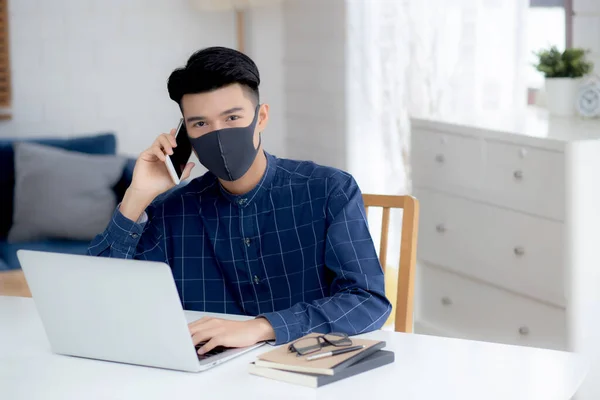 Young asian business man in face mask talking smartphone and work on laptop computer for protect covid-19, businessman quarantine work from home with notebook, social distancing and stay home.