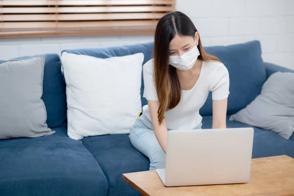 Young asian business woman in face mask work from home on laptop computer online to internet in living room, girl in medical mask and quarantine for pandemic of covid-19, stay home, new normal.