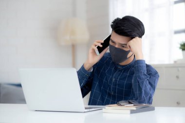 Yüz maskeli Asyalı genç bir iş adamı akıllı telefondan konuşuyor ve bilgisayar başında Coperid-19 'u korumak için çalışıyor. İş adamı karantina işini evden defter ve sosyal mesafeyle yapıyor ve evde kalıyor..