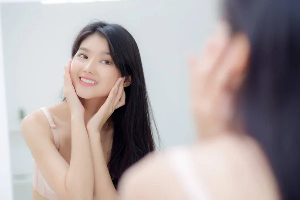 Beautiful young asian woman smiling look at mirror of checking face with skin care and cosmetic for rejuvenation and hygiene, beauty girl happy clean facial with cream or lotion and for health.