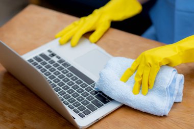 Evde pandemik covid-19 'u korumak için dizüstü bilgisayarı dezenfekte ve alkolle temizleyen Asyalı genç bir kadın, hijyen için eldiven temizleme defteri içindeki bir kız, sağlık konsepti.