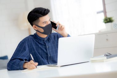 Yüz maskeli Asyalı genç bir iş adamı akıllı telefondan konuşuyor ve bilgisayarla covid-19 'u koruyor, iş adamı karantina işini evden yapıyor ve not defteri yazıyor, sosyal mesafe koyuyor ve evde kalıyor..