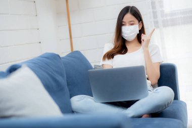 Yüz maskeli Asyalı genç bir iş kadını bilgisayardan internete, oradan oturma odasına, oradan tıbbi maskeli bir kız ve kovid-19 için karantinaya alma fikrini ve çalışmasını düşün..