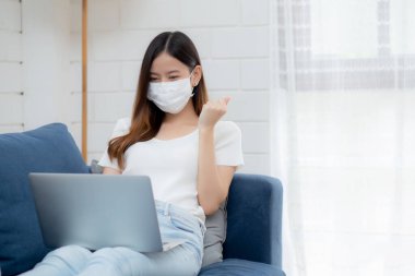 Yüz maskeli Asyalı genç bir iş kadını dizüstü bilgisayardan internet üzerinden oturma odasında internet başarısına, tıbbi maskeli bir kız ve kovid 19 için karantinaya alındı..