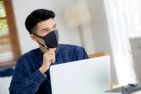 Young business man in face mask planning and working from home on laptop computer on desk for protection for outbreak of covid-19, male quarantine stay home using notebook on desk, new normal.