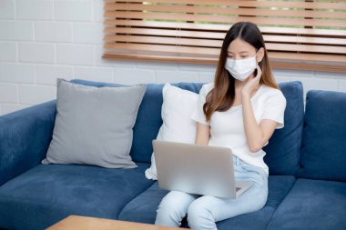 Yüz maskeli Asyalı genç bir iş kadını evde dizüstü bilgisayardan internete, oturma odasında internete, tıbbi maskeli bir kız ve covid-19 salgını için karantinaya alınıyor..