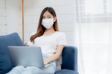 Yüz maskeli Asyalı genç bir iş kadını evde dizüstü bilgisayardan internete, oturma odasında internete, tıbbi maskeli bir kız ve covid-19 salgını için karantinaya alınıyor..