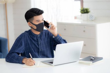 Yüz maskeli Asyalı genç bir iş adamı akıllı telefondan konuşuyor ve bilgisayarla covid-19 'u koruyor, iş adamı karantina işini evden yapıyor ve not defteri yazıyor, sosyal mesafe koyuyor ve evde kalıyor..