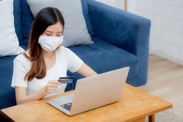 Young asian woman in face mask holding credit card shopping online with laptop computer buy and payment, girl using debit card purchase or transaction of finance, lifestyle and new normal concept.