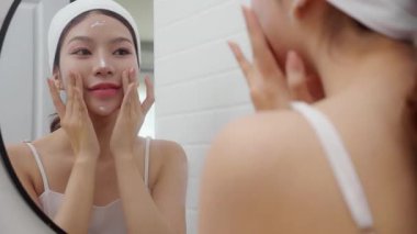 Genç ve güzel Asyalı bir kadın banyoda aynaya bakarken cildine krem sürüyor ya da losyon sürüyor. Tedavi için nemlendirici sürüyor. Yüz hatları, güzellik ve cilt bakımı için..