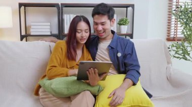 Genç Asyalı çift rahatlamak için tablete bakıyor. Oturma odasındaki kanepede rahatça oturuyorlar. Kanepede oturup tablet izliyorlar..