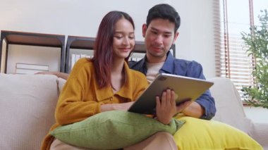 Genç Asyalı çift rahatlamak için tablete bakıyor. Oturma odasındaki kanepede rahatça oturuyorlar. Kanepede oturup tablet izliyorlar..