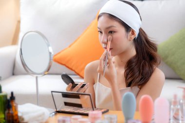 Güzel, mutlu, Asyalı bir kadın oturma odasında aynaya bakarak makyaj yapıyor. Güzel bir kadın makyaj yapıyor. Cazibe ve zarafet için yüz makyajı yapıyor..