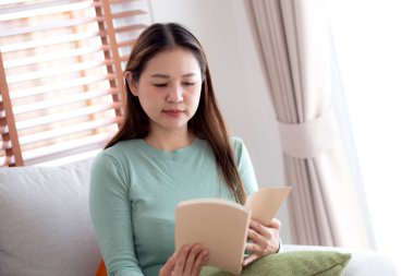 Genç Asyalı bir kadın, yüz ifadesiyle kitap okuyor. Oturma odasındaki koltukta oturuyor, kadın rahatlıyor ve kanepeye oturuyor. Edebiyat, etkinlik ve hobi, yaşam tarzı ve rahatlama okuyor..