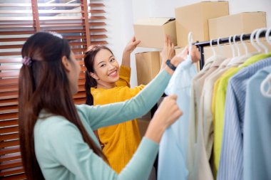Hazırlık teslimatı için mağazada kıyafet düzenleyen iki genç Asyalı kadın, satıcı seçici giysiler, perakende satış için seçici giysiler, teslimat için mal, küçük işletme.