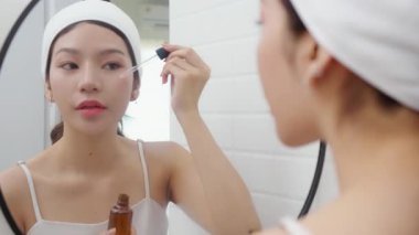 Güzel Asyalı genç bir kadın, yüz aynasına gençleştirme serumu uyguluyor. Güzellik kadını cilt, cilt bakımı, cilt bakımı, cilt bakımı, makyaj, kozmetik ve makyaj için kolajen uyguluyor..