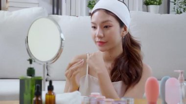 Güzel, genç Asyalı bir kadın, evde aynaya bakarken krem veya losyon sürüyor. Mutlu, güzel bir kadın, ellerinde gençleştirme, cilt bakımı ve sağlık hizmetleri için cilt bakımı yapıyor..