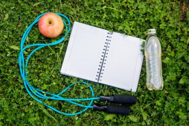 Bir şişe su, defter, elma ve taze yeşil çimenlerin üzerinde atlama ipi. Üstten görünüm, kopya alanı.