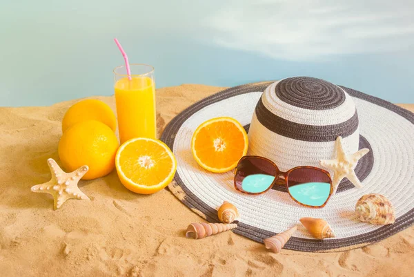 Glass of orange juice, hat, sunglasses and seashells on sand beach on blue backdrop