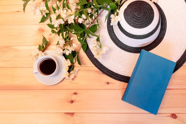 Summer hat, cup of coffee, book and jasmine flowers on wooden background. Top view. Summer rest concept.