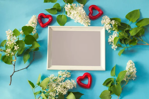 White frame, flowers of white lilac and red hearts on blue background.