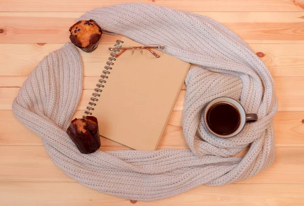 Taccuino vuoto, tazza di caffè caldo avvolto in sciarpa, bicchieri e muffin su sfondo di legno . — Foto Stock