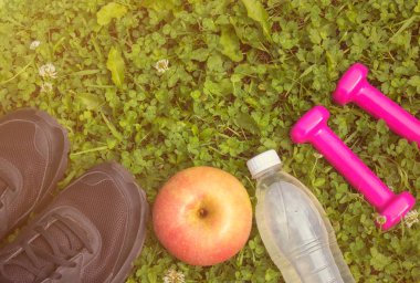 Siyah spor ayakkabı, pembe dambıl, şişe suyu ve elma yeşil çimenlerin üzerinde.