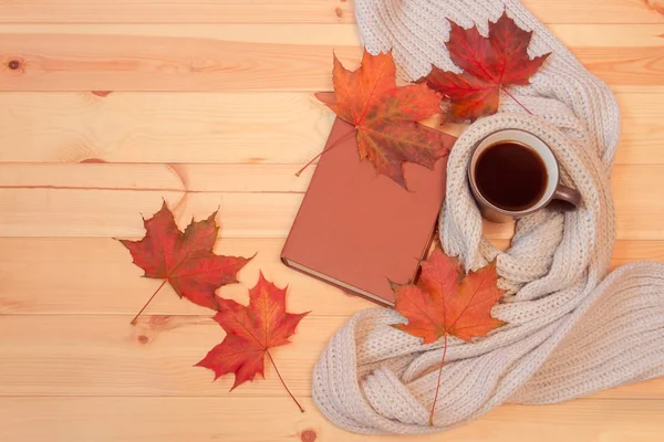 Hrnek horké kávy, podzimní listy, kniha a šátek na dřevěné pozadí. — Stock fotografie