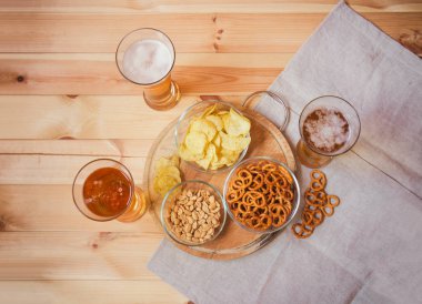 Ahşap masada patates cipsi, tuzlu kraker ve yer fıstığı ile hafif bira bardak. 