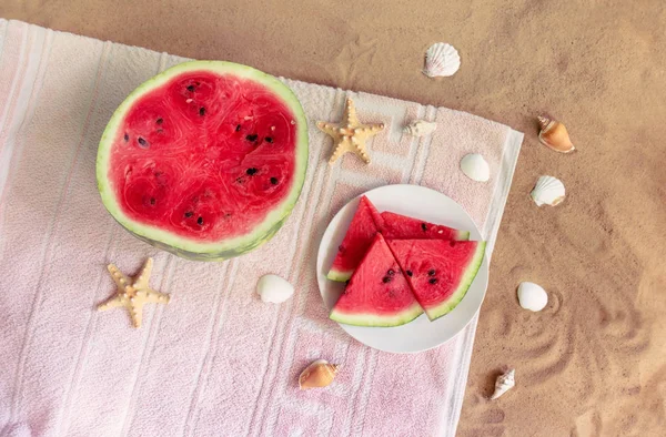 Metade de melancia, prato com fatias de melancia, estrelas do mar e conchas na toalha na praia de areia . — Fotografia de Stock