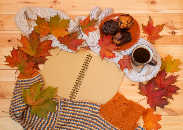 Cuaderno vacío, taza de café caliente, hojas de arce de otoño, magdalenas y suéter casero caliente en la superficie de madera . — Foto de Stock