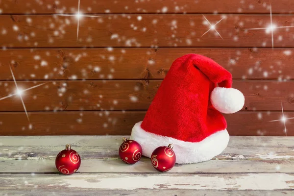 Fluffy Santa hat with christmas balls on aged wooden table near wooden wall. — Stock Photo, Image