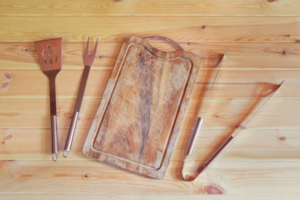Άδειο ξύλινο τραπέζι κοπής, πιρούνι για το κρέας, σπάτουλα και λαβίδες κουζίνας σε ρουστίκ ξύλινο φόντο. — Φωτογραφία Αρχείου