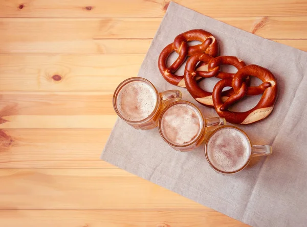 Tres tazas de cerveza y pretzels suaves en la mesa de madera. Vista superior, espacio de copia — Foto de Stock