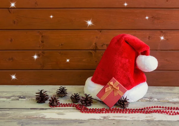 Fuffy Santa hat with pine cones and christmas gift on aged wooden table near wooden wall. — Stock Photo, Image