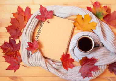 Bir kupa sıcak kahve, boş defter, eşarp ve ahşap arka planda renkli sonbahar akçaağaç yaprakları. Üst görünüm, alanı kopyala.