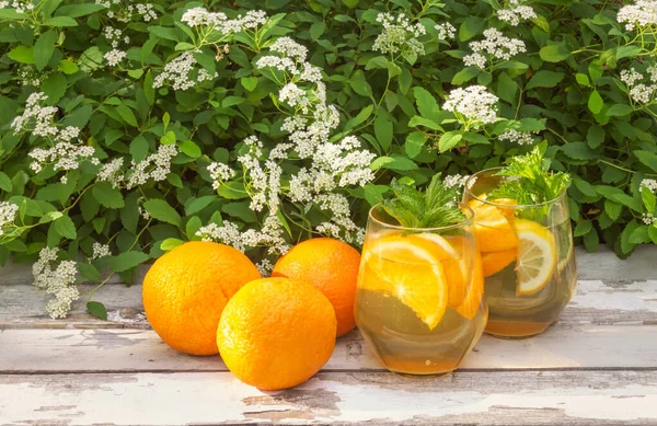 Summer orange lemonade in glasses and fresh oranges near on rustic wooden table on fresh white Spiraea flowers background