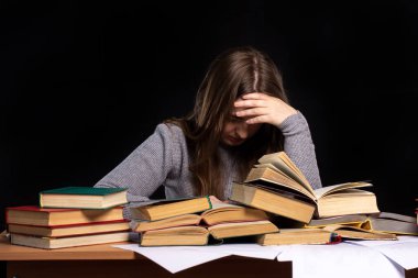Genç bir kız masasında oturuyor ve elini yüzünü kapatarak kitap okuyor. Eğitimde zorluk. Uzaktan öğrenme. Arkaplanda siyah duvar.