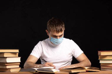 Tıbbi maskeli bir adam kitaplarla dolu bir masada oturuyor. Belgelerin incelenmesine tepeden bak. Karantina sırasında çalış. Uzaktan çalışmanın zorlukları. Öğretmen uzaklık dersi için hazırlanıyor..