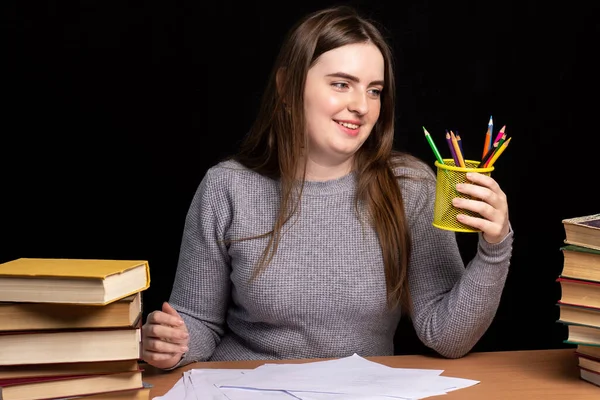a young girl sits at a table with books and documents in her hand holds a glass with pencils. Remote education. the teacher is preparing for the lesson. office worker in a working environment. Black background