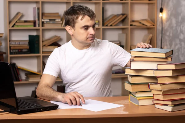 young man looks at a stack of books on the background of a bookcase. a laptop is standing nearby. student is preparing for the exam. the teacher checks homework. An office employee is studying reports.
