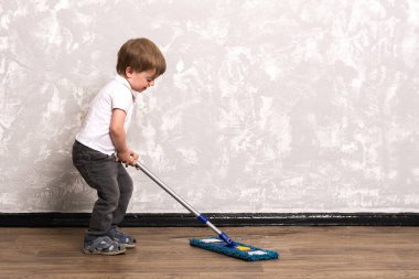 Küçük çocuk gri bir doku duvarına karşı yerleri paspaslıyor. Üç yaşındaki bir çocuk, ebeveynlerinin evde bahar temizliği yapmasına yardım ediyor..