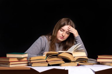 Genç bir kız öğrenci, elinde kitaplarla bir masada oturuyor. Kafasını siyah bir duvarın arka planına yaslıyor. konsept ve evde eğitim zorlukları. Yorgunum. Belgelere bak..