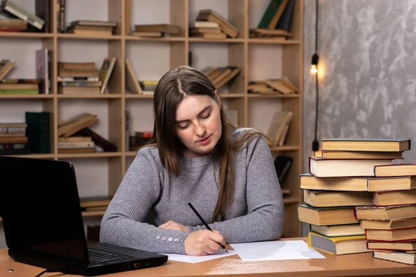 若い女子学生が授業の準備をしています オフィスワーカーが文書で書いてる ノートパソコンはテーブルの上にあり背景には本棚があります 仕事中のフリーランスは — ストック写真