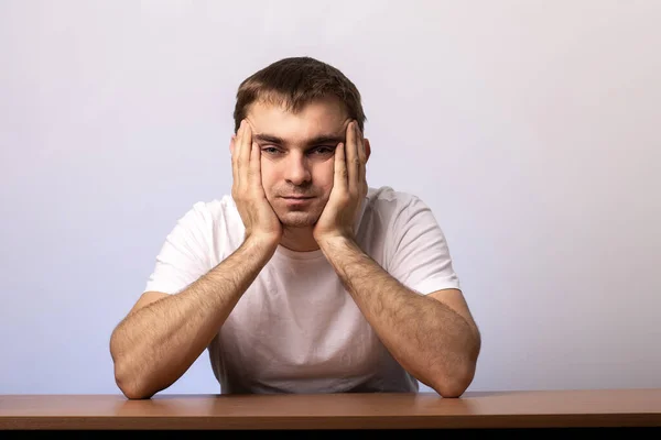 stock image a young man sits at a table against a wall background holding his face with his hands.