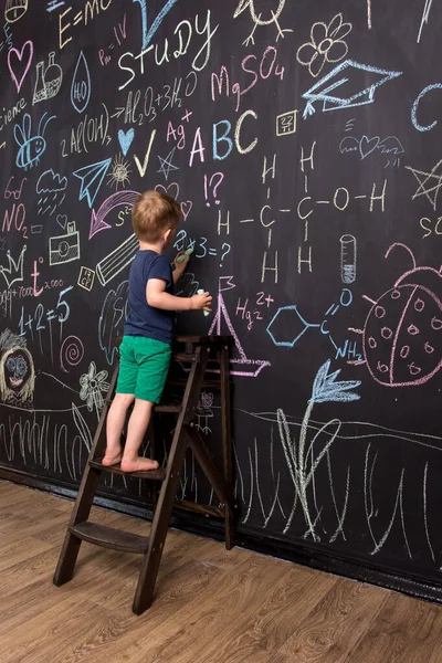 小男孩用彩色粉笔在一块大黑板上画画 有儿童图画和公式的黑墙 垂直摄影 — 图库照片