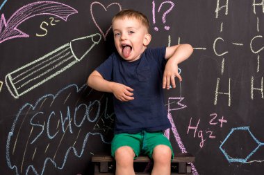 Üç yaşındaki küçük bir çocuk büyük bir tahta duvarın yanında duruyor ve renkli tebeşirle çizimler yapıyor. Avrupa görünüşü. Çocuk şımarır ve dil gösterir.